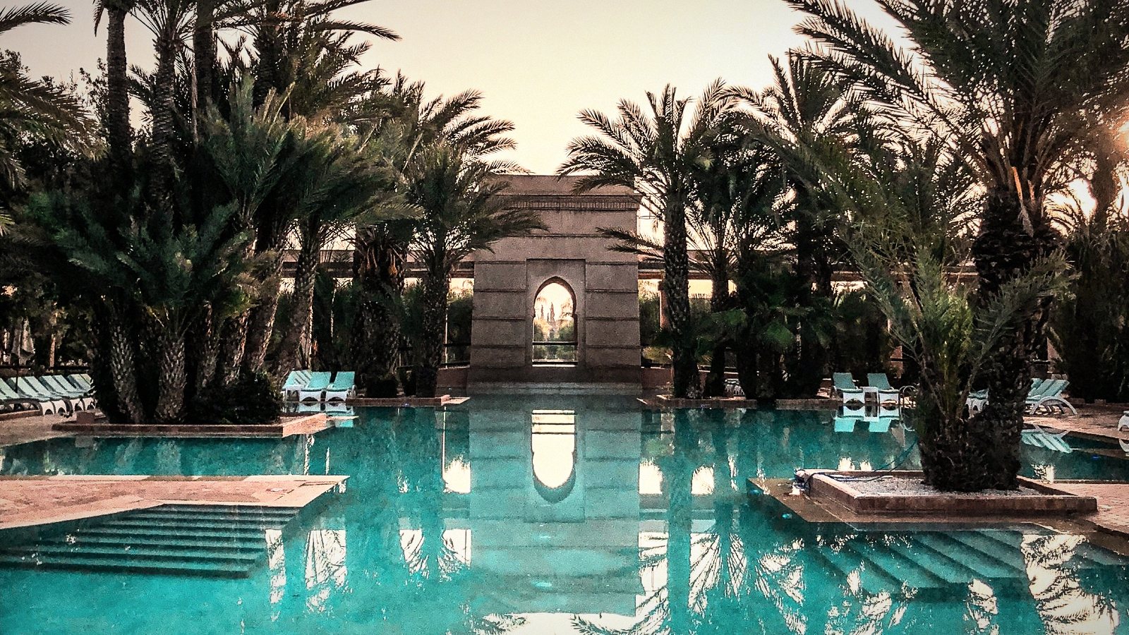 A long reflecting pool flanked by date palms, a stone pavilion mirrored at the far end