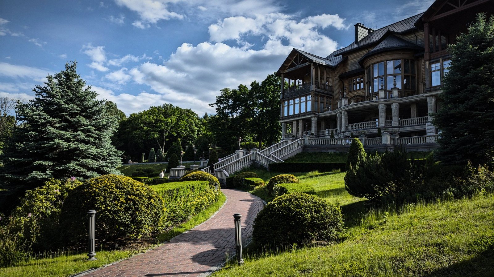 A large house on a landscaped hillside, a brick path climbing between clipped hedges and pines
